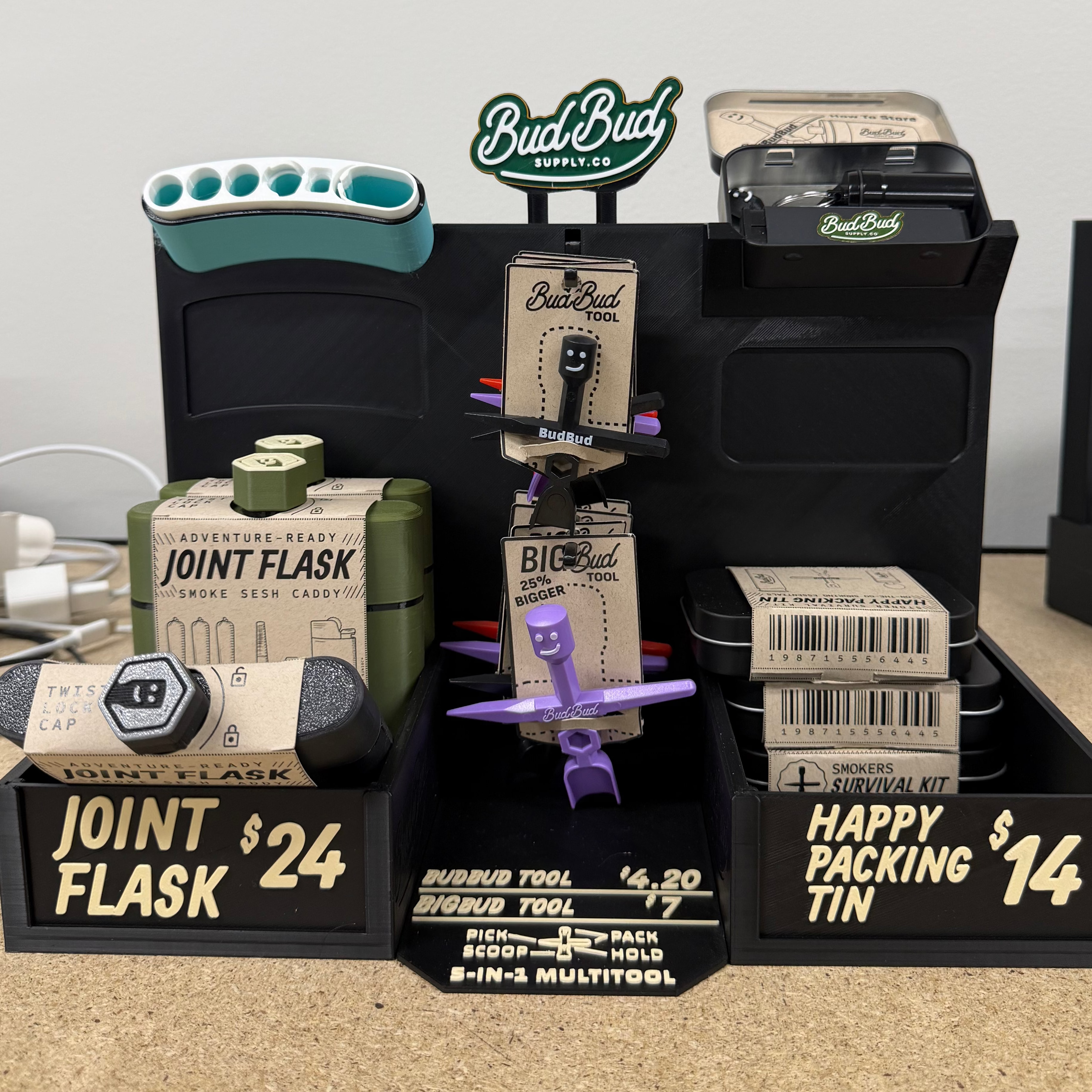 A tabletop display of various joint distillations and smoking accessories on a beige carpet against a light gray wall. The products include a BudBud Joint Distillation machine, a Joint Flask, a Happy Packing Tin, and other smoking tools in black, green, and purple packaging.